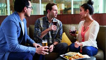 Three people seated on a couch, holding glasses of beer, with a plate of snacks on a table in front of them.