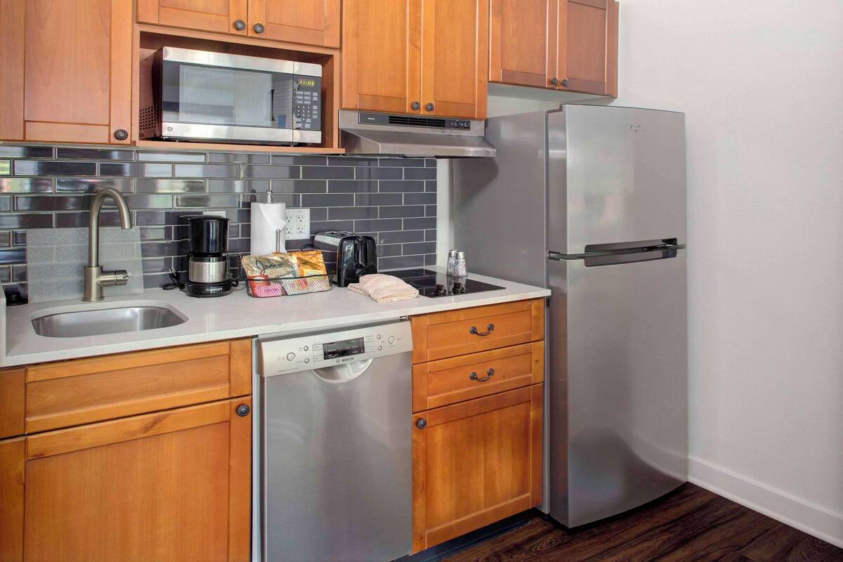 Cocina compacta con armarios de madera, un refrigerador de acero inoxidable, microondas, lavavajillas y cafetera.