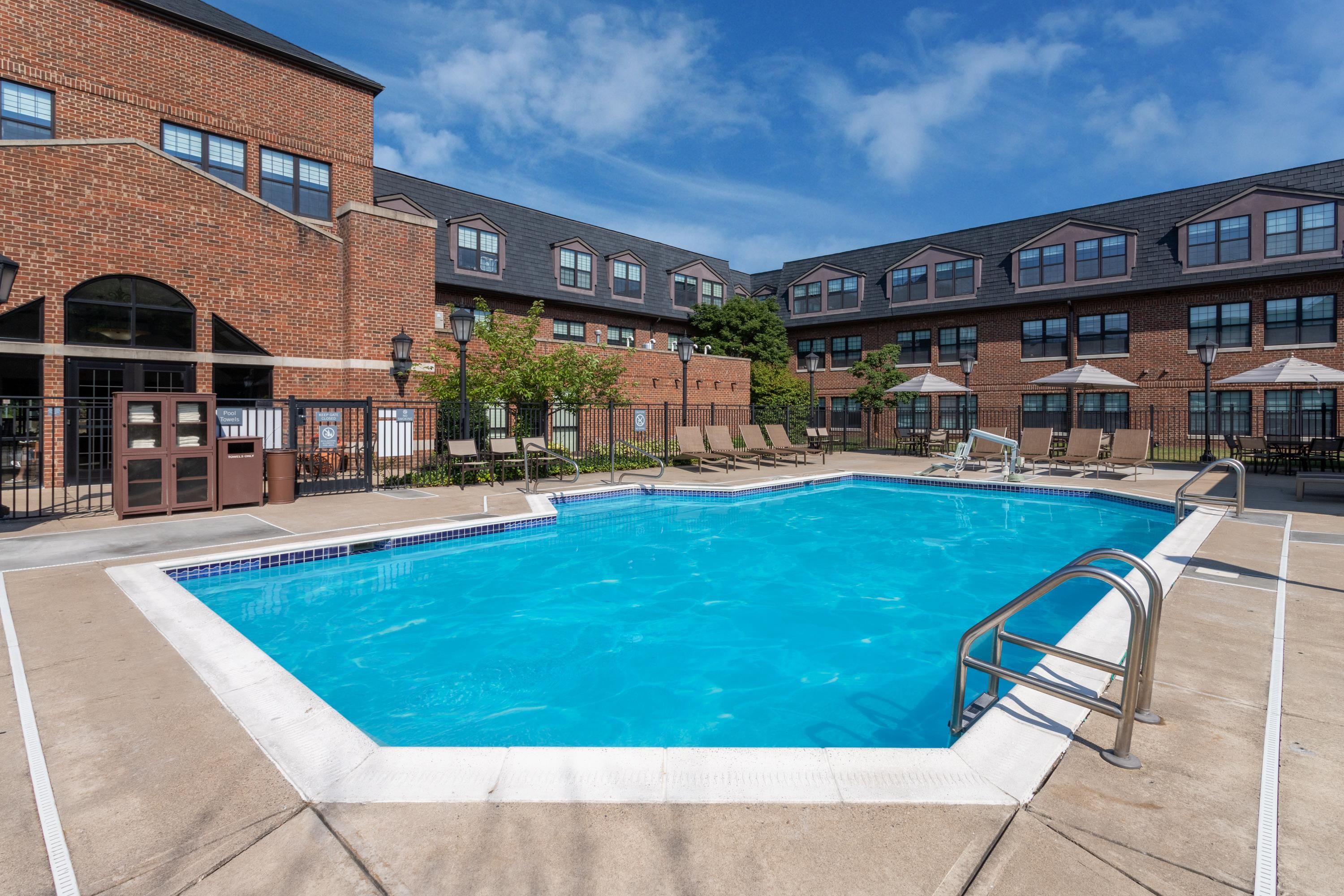 Une piscine rectangulaire bordée de carreaux, entourée d’un bâtiment en briques doté de nombreuses fenêtres et d’un espace détente couvert avec des parasols.