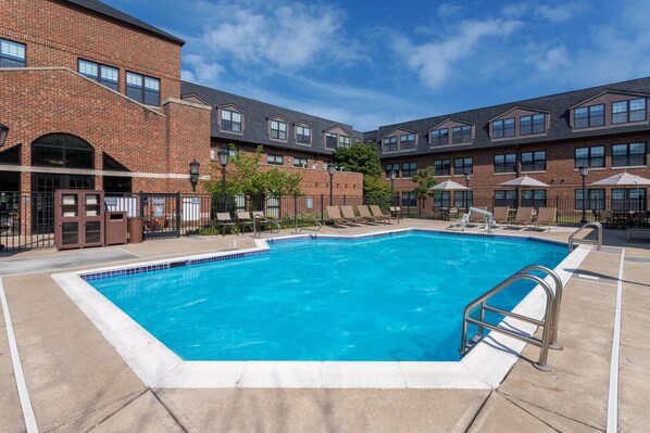 A rectangular pool with a tiled edge, surrounded by a brick building with multiple windows and a covered seating area with umbrellas.