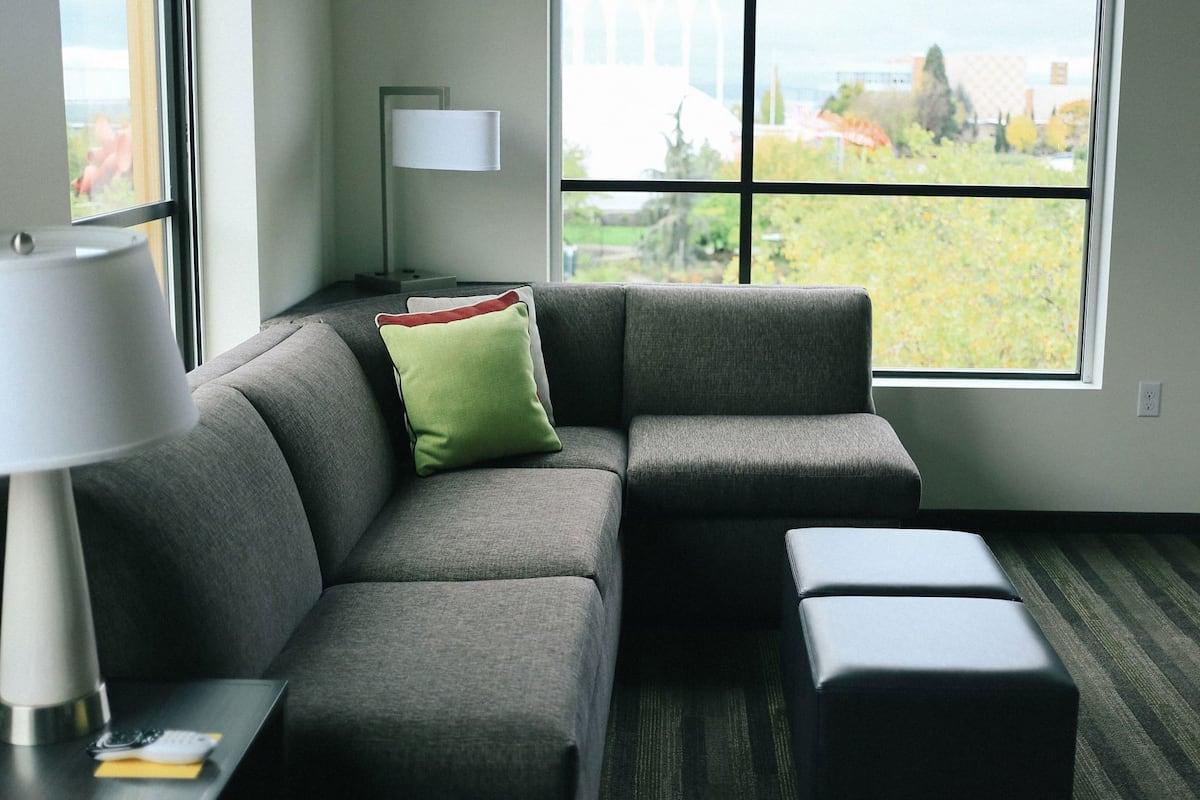 Una sala moderna con un sofá seccional gris, un cojín verde, una mesita de noche con una lámpara y vistas a la vegetación a través de amplias ventanas.