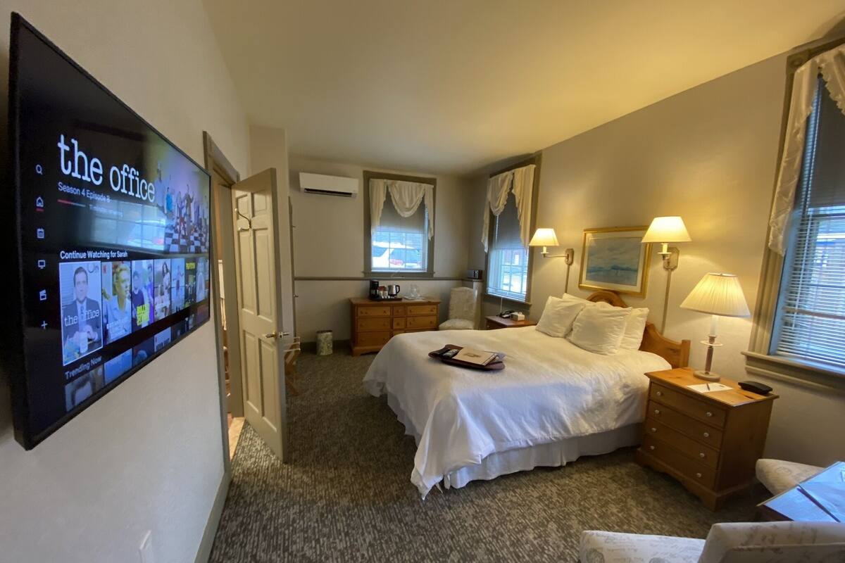 A hotel room with a bed, bedside tables, a TV mounted on the wall, a dresser, and a window with curtains.