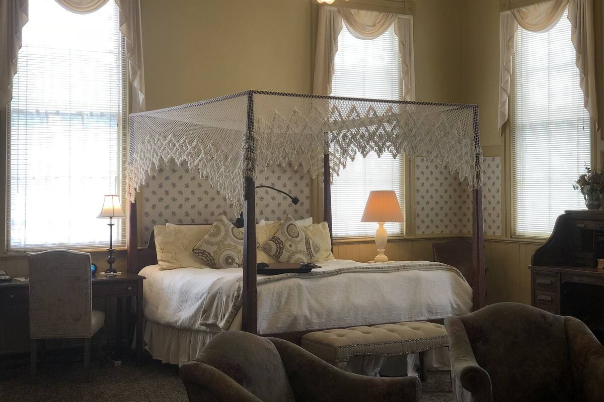A four-poster bed with a canopy, two bedside lamps, a desk with a chair, and a television on a stand.