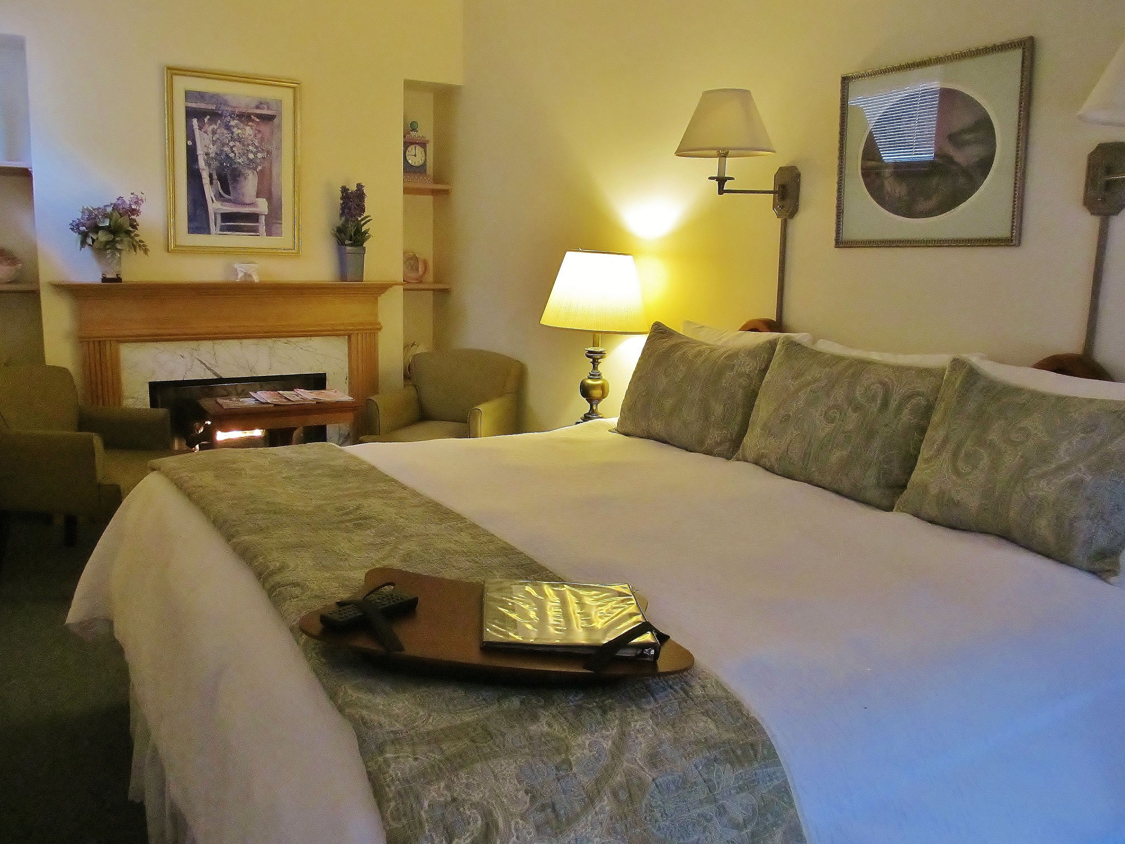 A hotel room with a large bed, a desk with a lamp, a chair, a fireplace, and a clock on the wall.