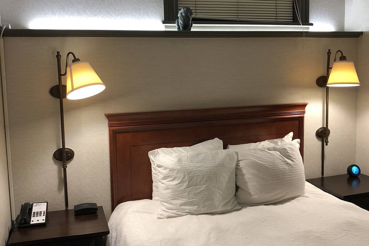 A hotel room with a bed, two bedside lamps, a nightstand with a phone, a clock, and an air conditioning unit.