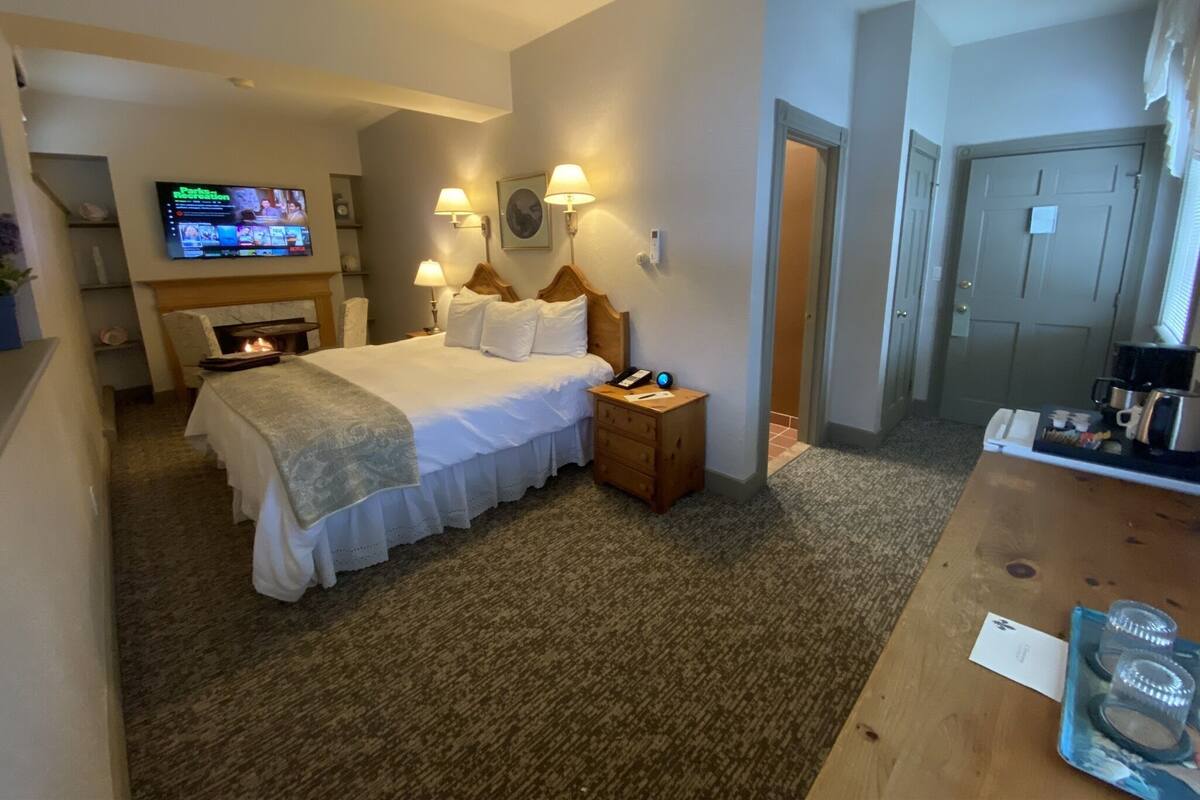 A hotel room with a bed, bedside lamps, a TV, a desk with a coffee maker, and a door.