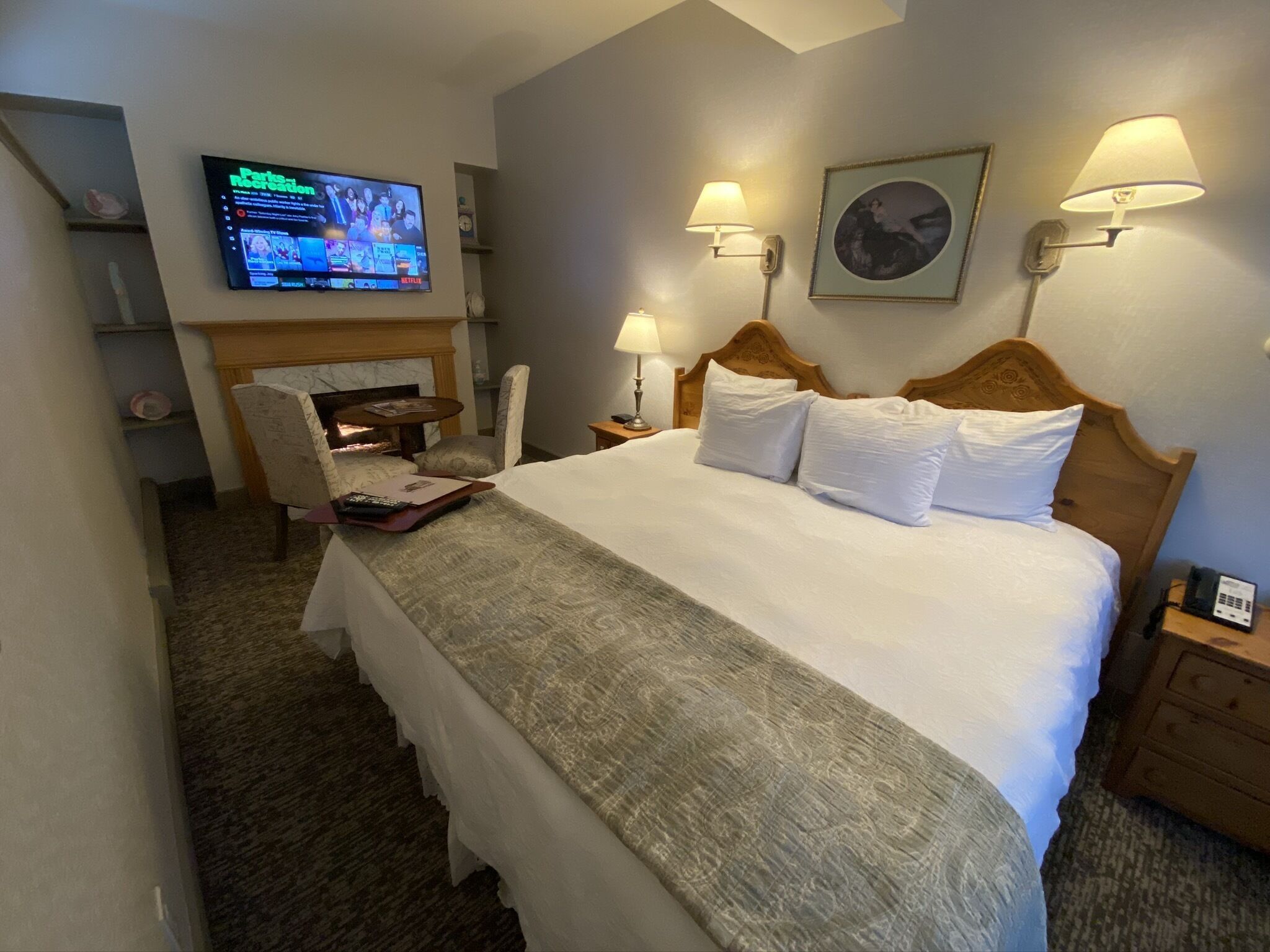 Habitación de hotel con cama, lámparas de noche, televisor montado en la pared, chimenea, una silla y una mesita.