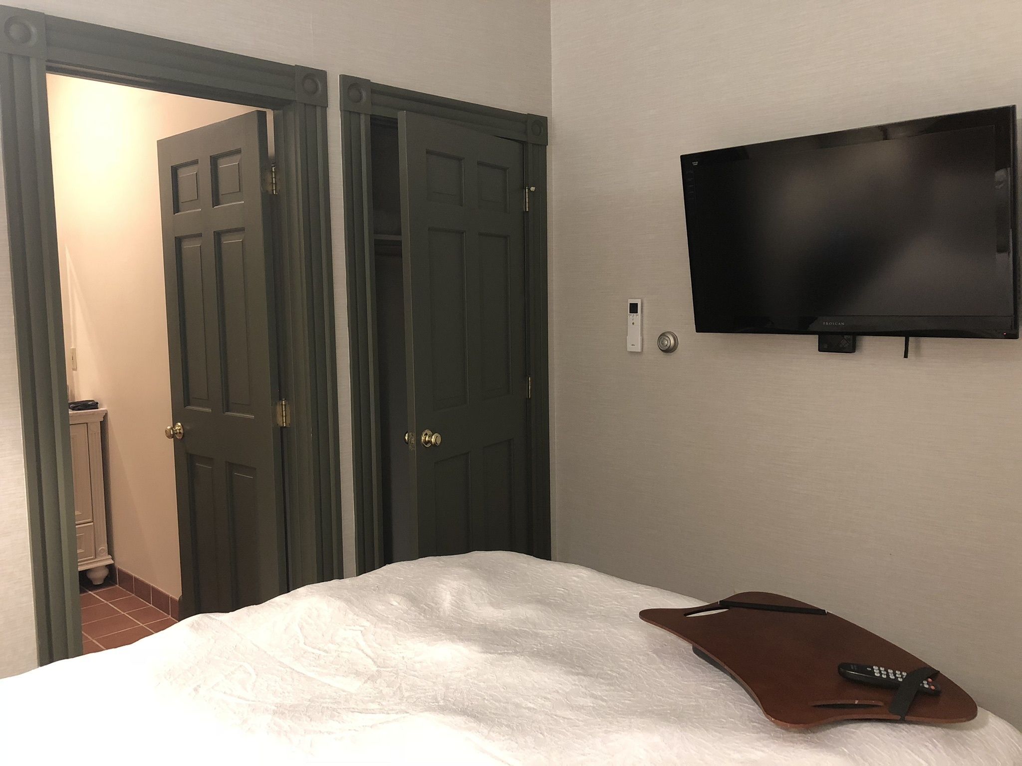 A hotel room with a bed, a remote control, a television mounted on the wall, and two doors.