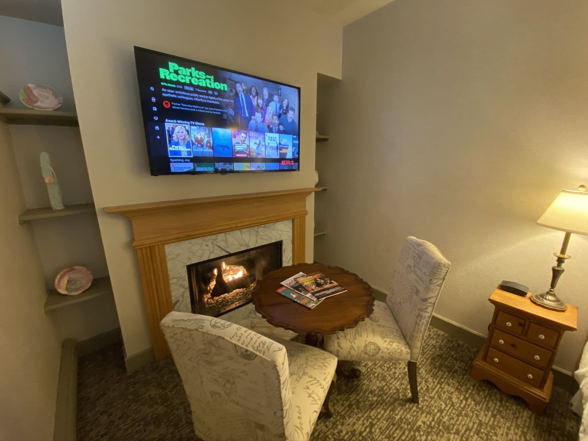 Hotelkamer met een aan de muur bevestigde televisie waarop een programma over parken en recreatie wordt vertoond. Er is een open haard met een schoorsteenmantel, een ronde houten tafel met tijdschriften, twee gestoffeerde stoelen en een nachtkastje met een lamp.