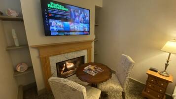 Hotelkamer met een aan de muur bevestigde televisie waarop een programma over parken en recreatie wordt vertoond. Er is een open haard met een schoorsteenmantel, een ronde houten tafel met tijdschriften, twee gestoffeerde stoelen en een nachtkastje met een lamp.
