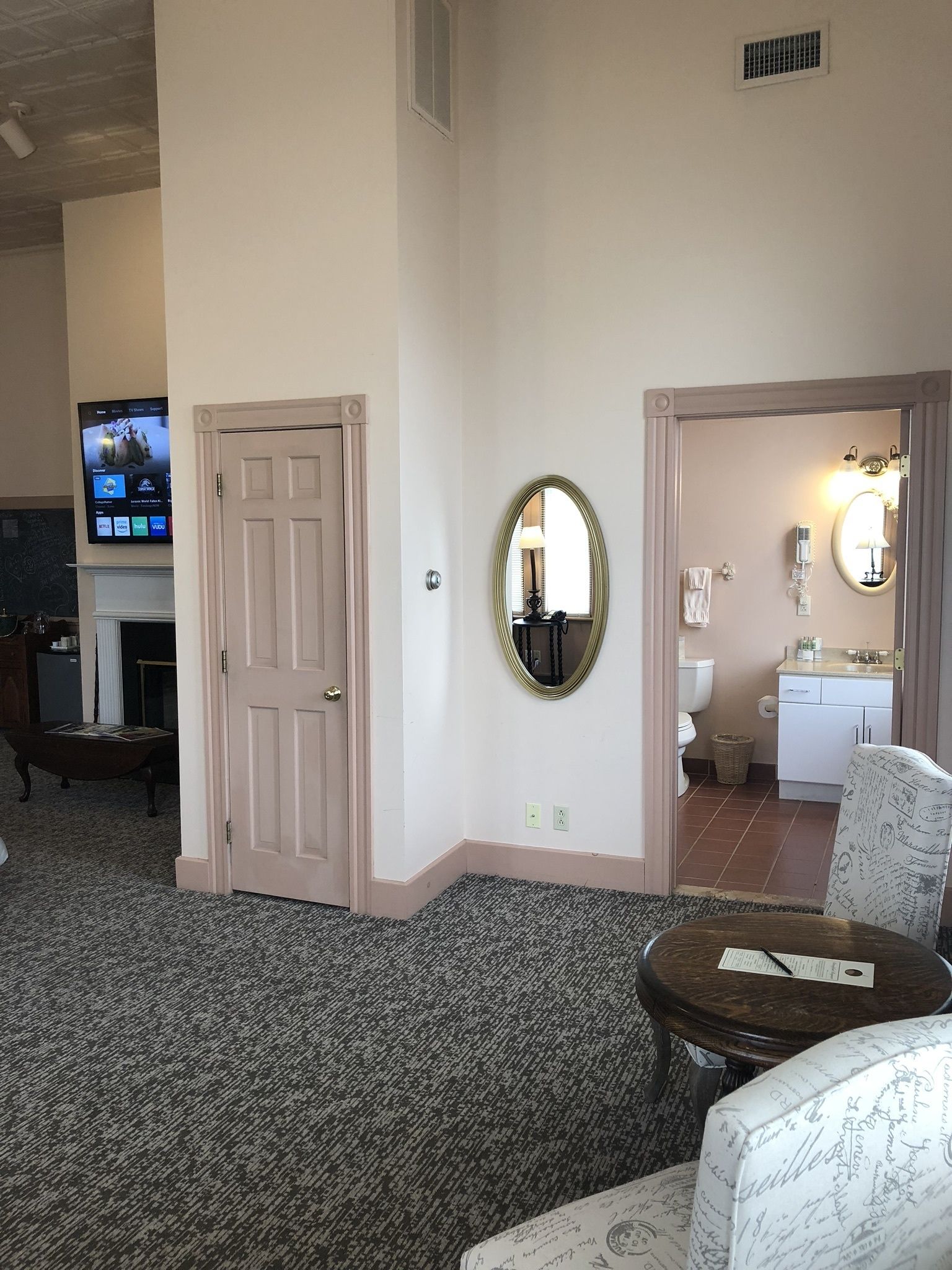 A room with a fireplace, a television mounted on the wall, a mirror, and a chair with a patterned cushion.