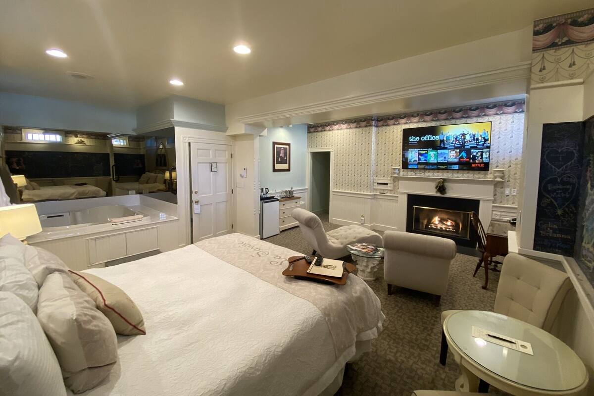 A hotel room with a large bed, a TV mounted above a fireplace, a desk, and a seating area with chairs.