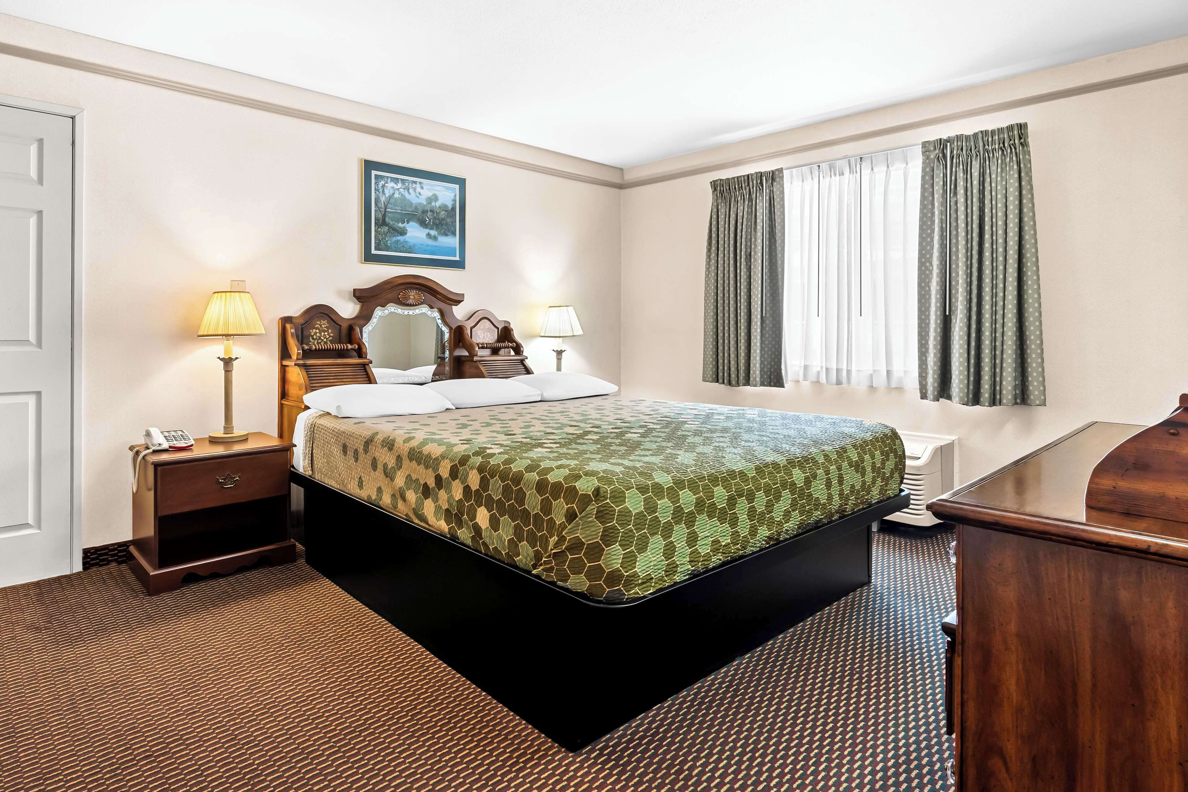 Quarto de hotel com cama grande, duas mesas de cabeceira com abajures, cabeceira de madeira, um quadro na parede e uma janela com cortinas.