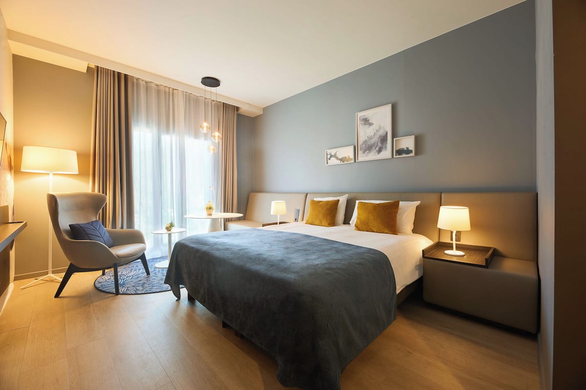 A hotel room with a bed, a chair, a bedside table, a lamp, and a window with curtains.