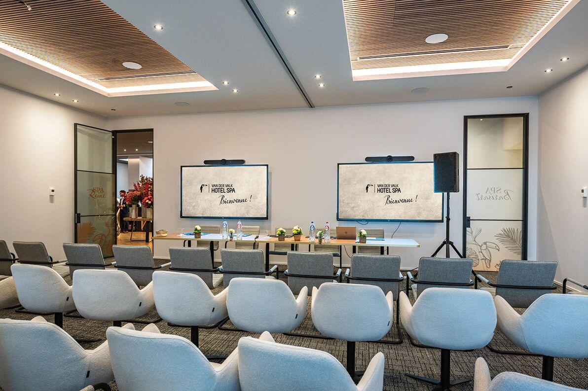 Un salón de conferencias con filas de sillas, una mesa con computadoras portátiles y dos pantallas mostrando 'Welcome to Van der Valk Hotel Spa'.
