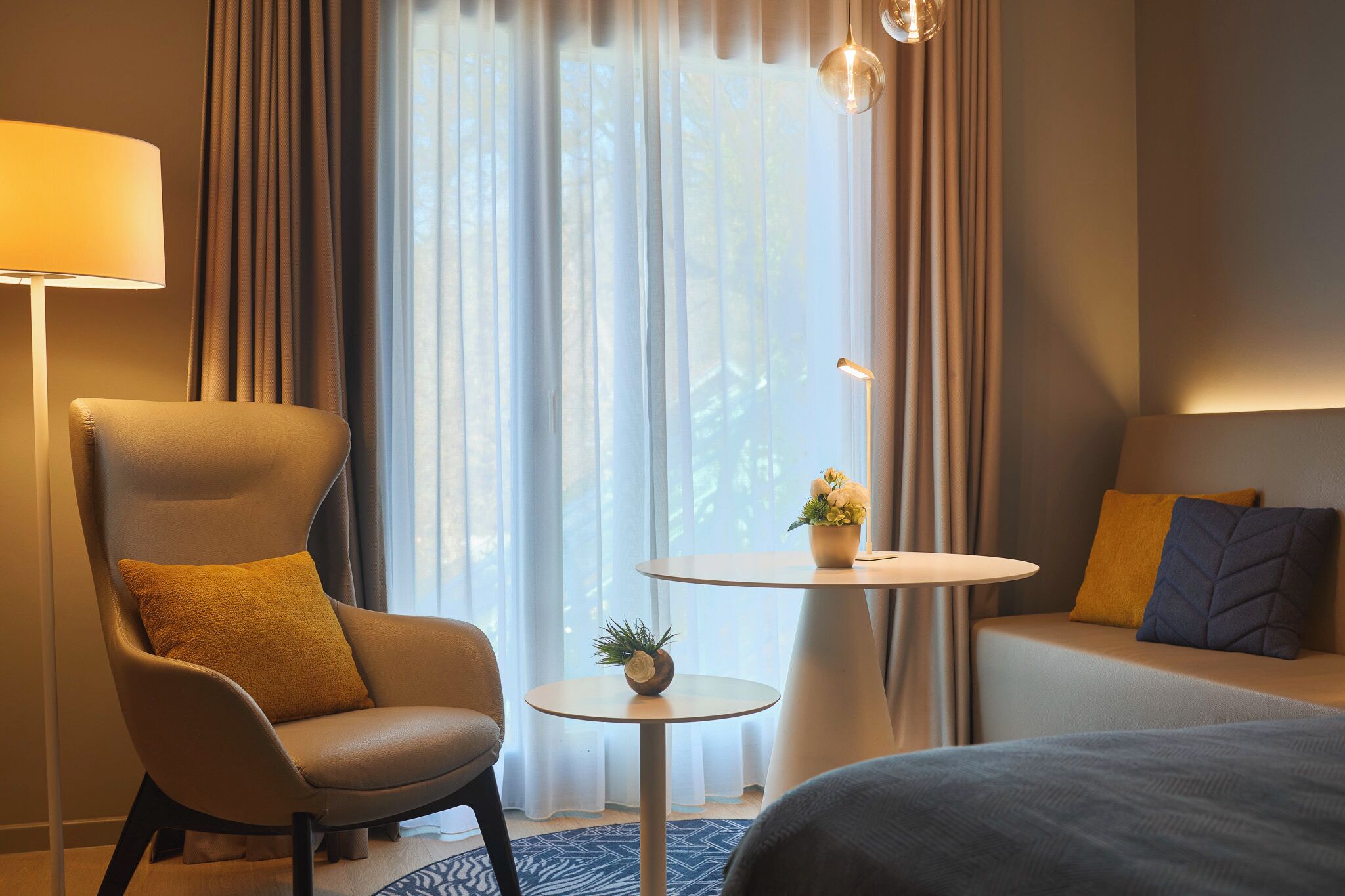A hotel room with a large window, a round table with a plant, a sofa with cushions, and a chair with a cushion.