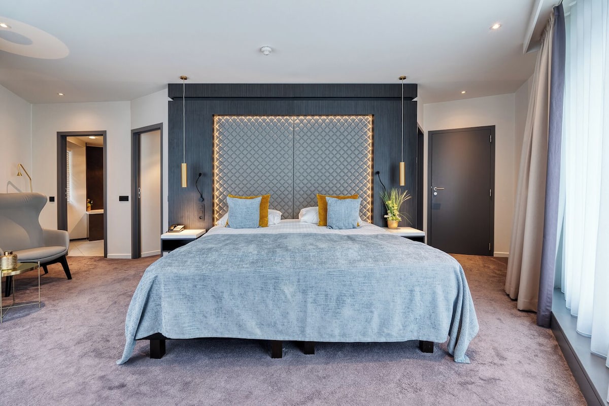 A modern hotel bedroom with a large bed, a headboard with a geometric pattern, a bedside table with a lamp, and a view of the hallway.