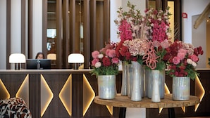 A reception desk with a computer, a wooden table with vases of flowers, and a patterned chair. - Van der Valk Hotel Spa (Spa)