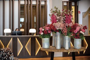 A reception desk with a computer, a wooden table with vases of flowers, and a patterned chair. - Van der Valk Hotel Spa (Spa)