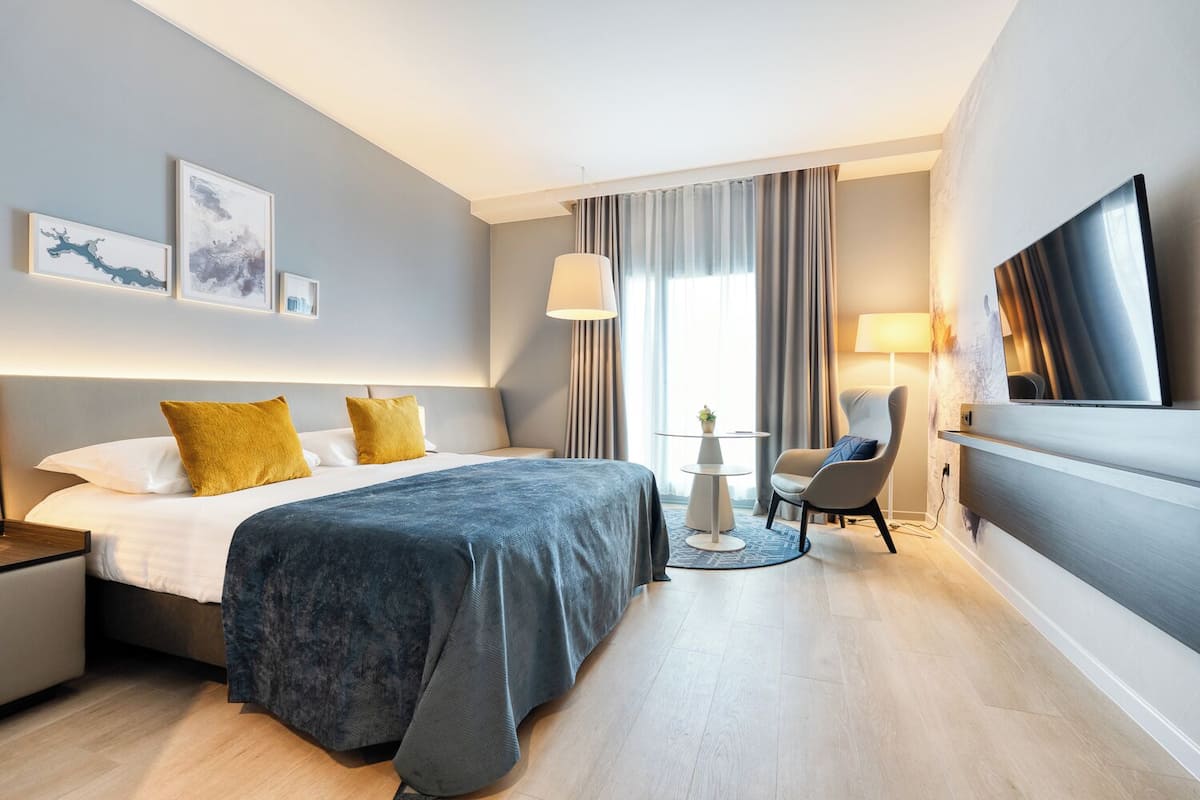 A modern hotel room with a bed, bedside tables, a desk, a chair, a TV, and artwork on the walls.