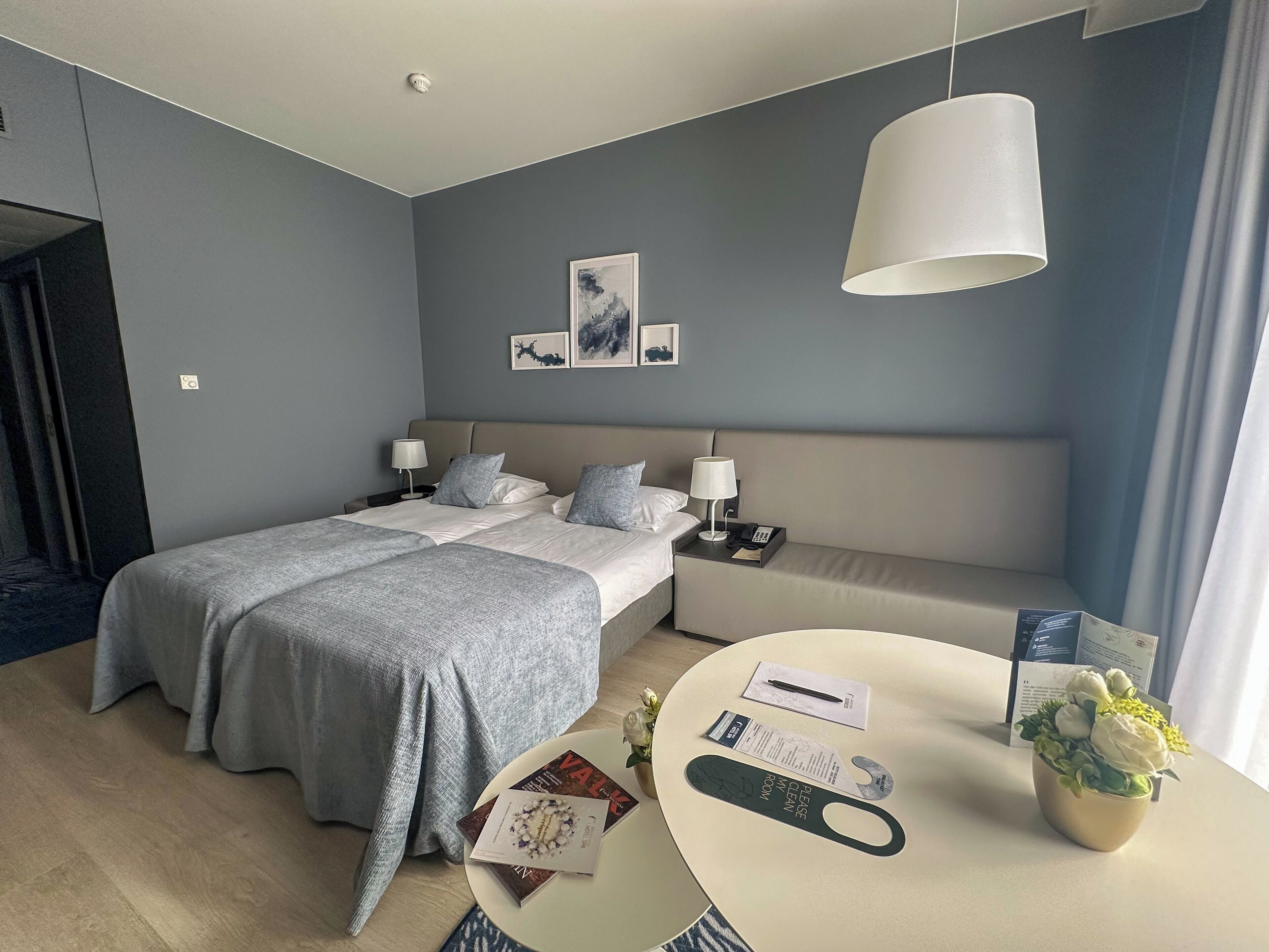 Una habitación de hotel moderna con una cama, mesitas de noche, una mesa redonda con un florero y una lámpara de pared.