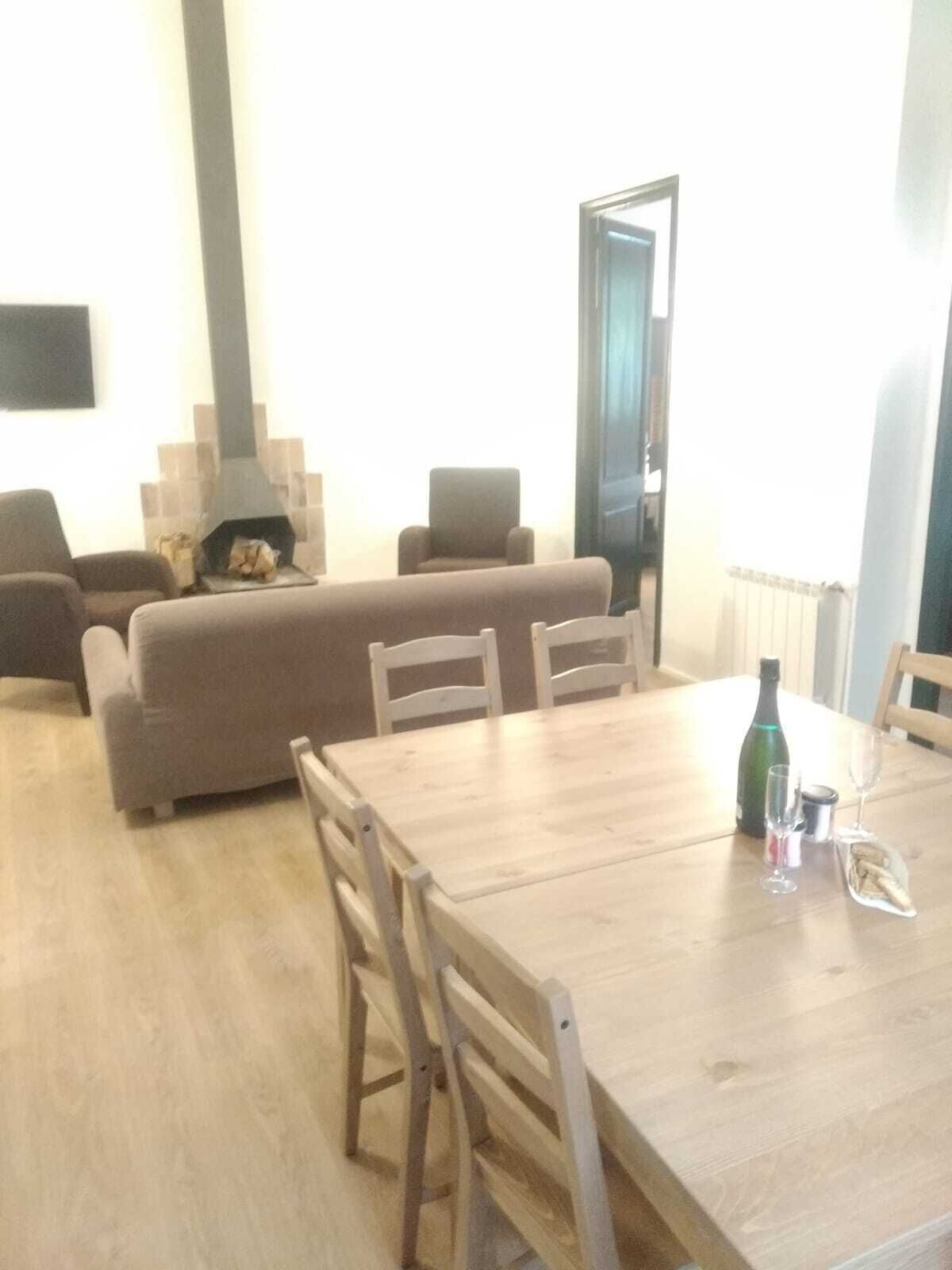 Una sala de estar con sofá, sillas y una mesa de comedor de madera con una botella, un vaso y aperitivos.