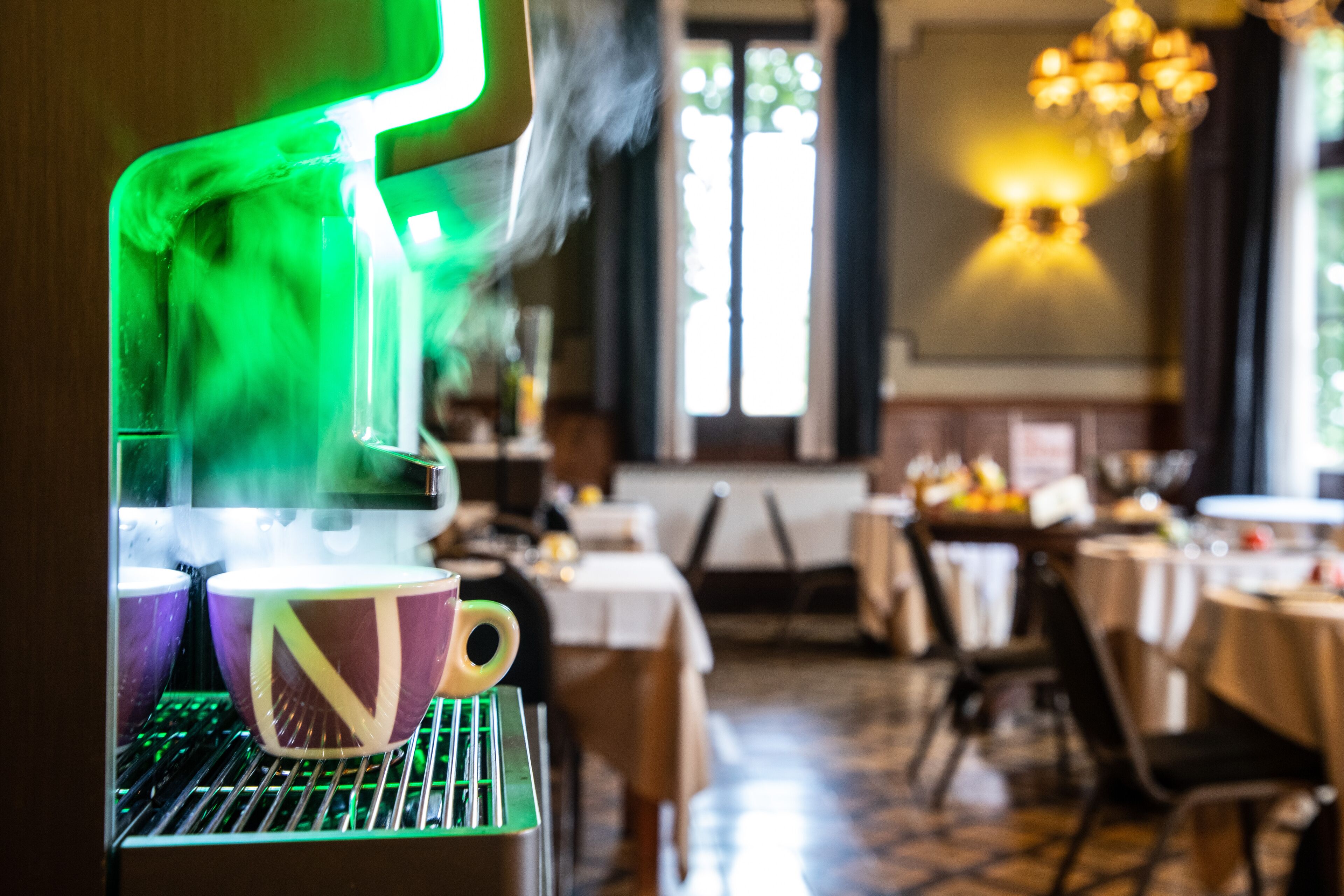 Een koffieapparaat schenkt hete koffie in bekers in een restaurant met tafels en stoelen.