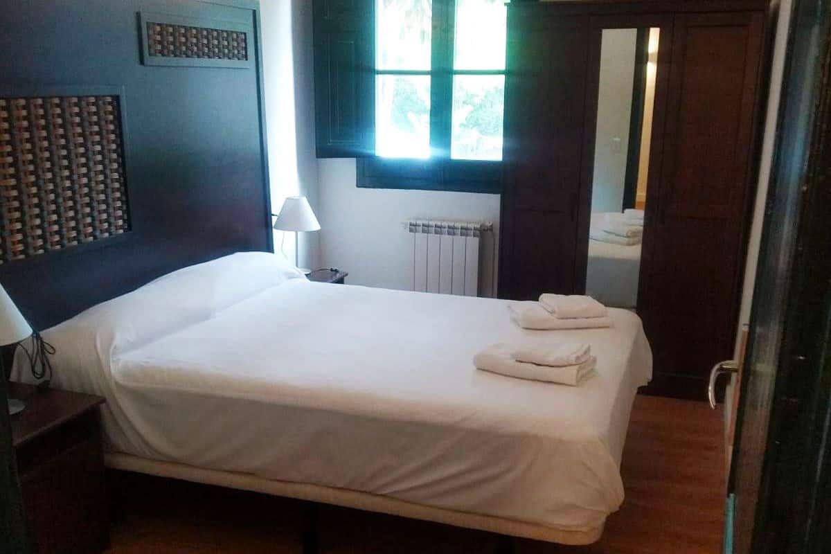 Een hotelkamer met een groot bed, een hoofdeinde met een ruitpatroon, een nachtkastje met een lamp, een raam met luiken en een radiator.