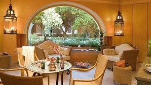 Un rincón acogedor con una mesa redonda, sillas de mimbre y un sofá. Un amplio ventanal de arco ofrece vistas al exterior, con vegetación.
