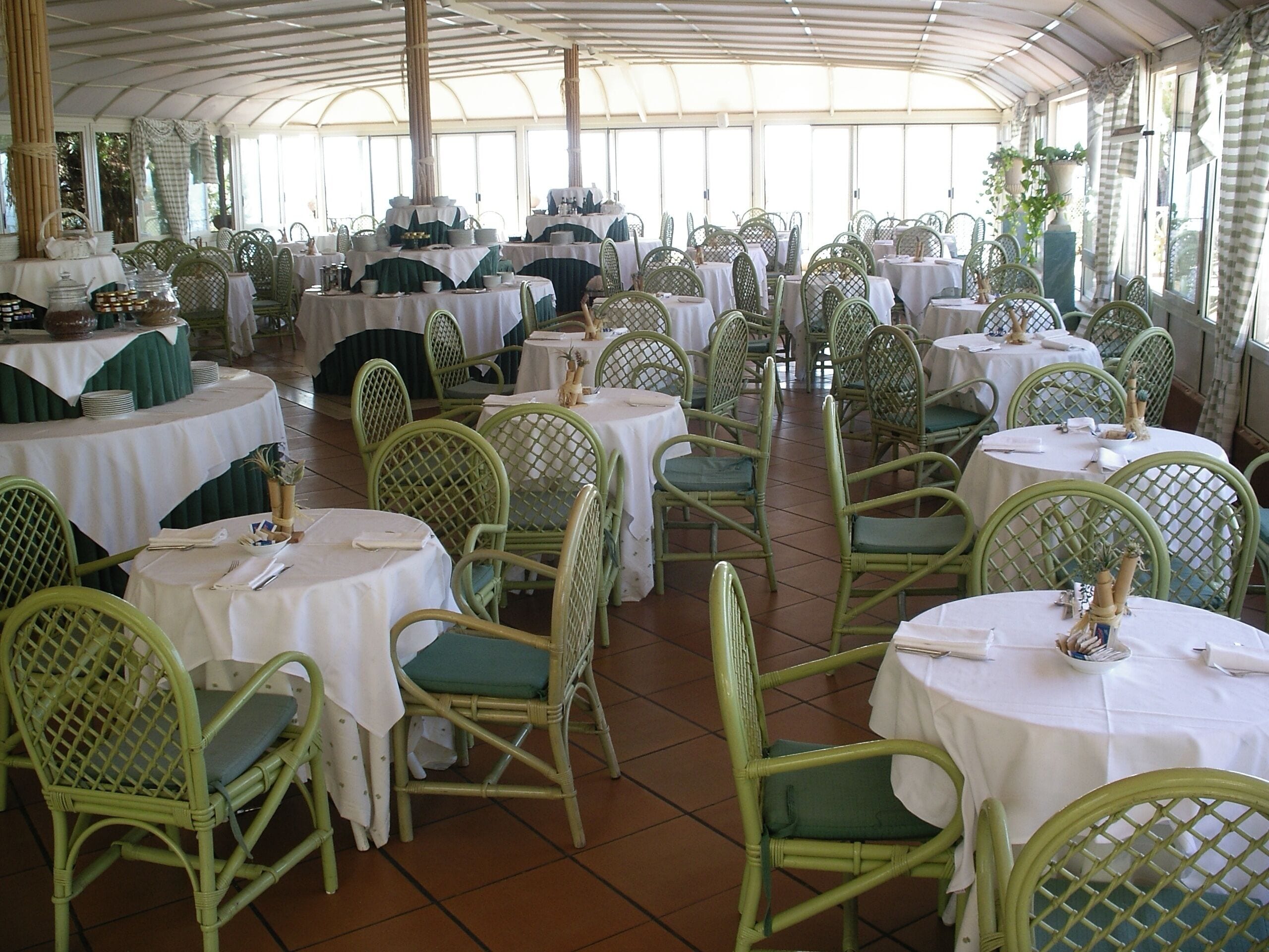 A covered dining area with round tables and green wicker chairs, featuring white tablecloths and potted plants.