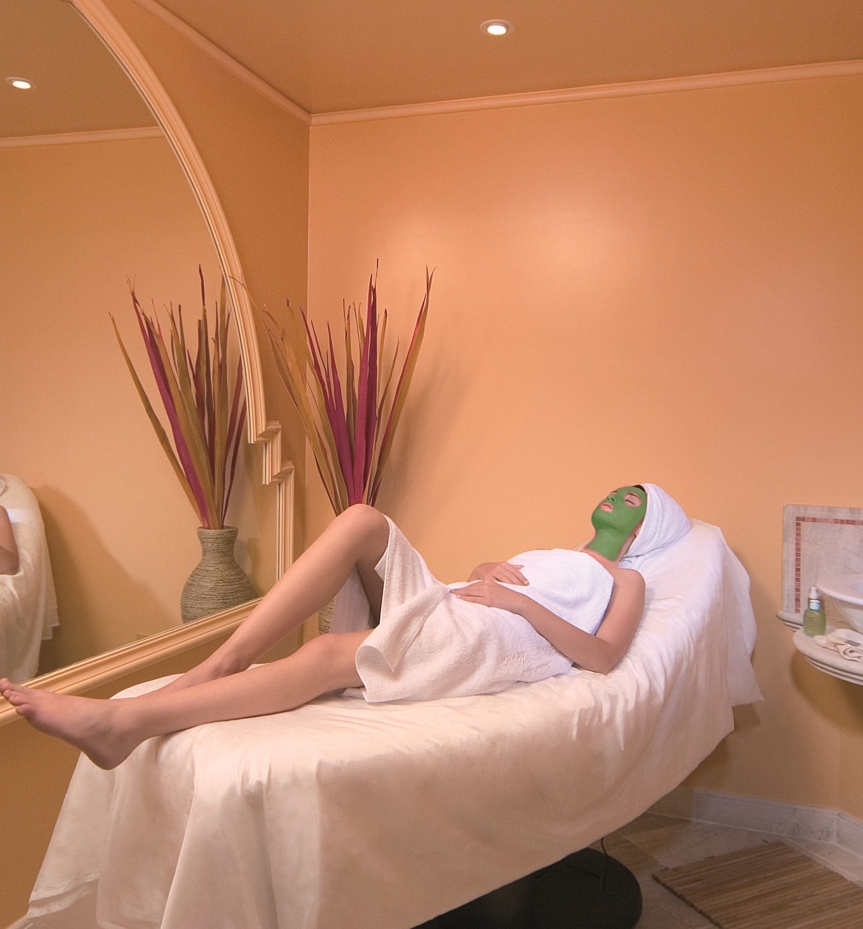 A person lying on a bed in a room with a mirror, a vase with tall plants, and a small table with a bottle and a container.