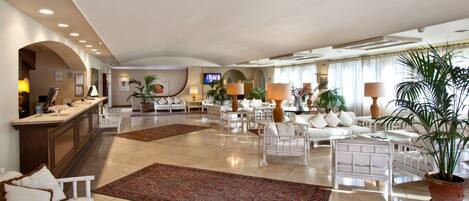A spacious hotel lobby with a reception desk, seating areas, and a television screen.