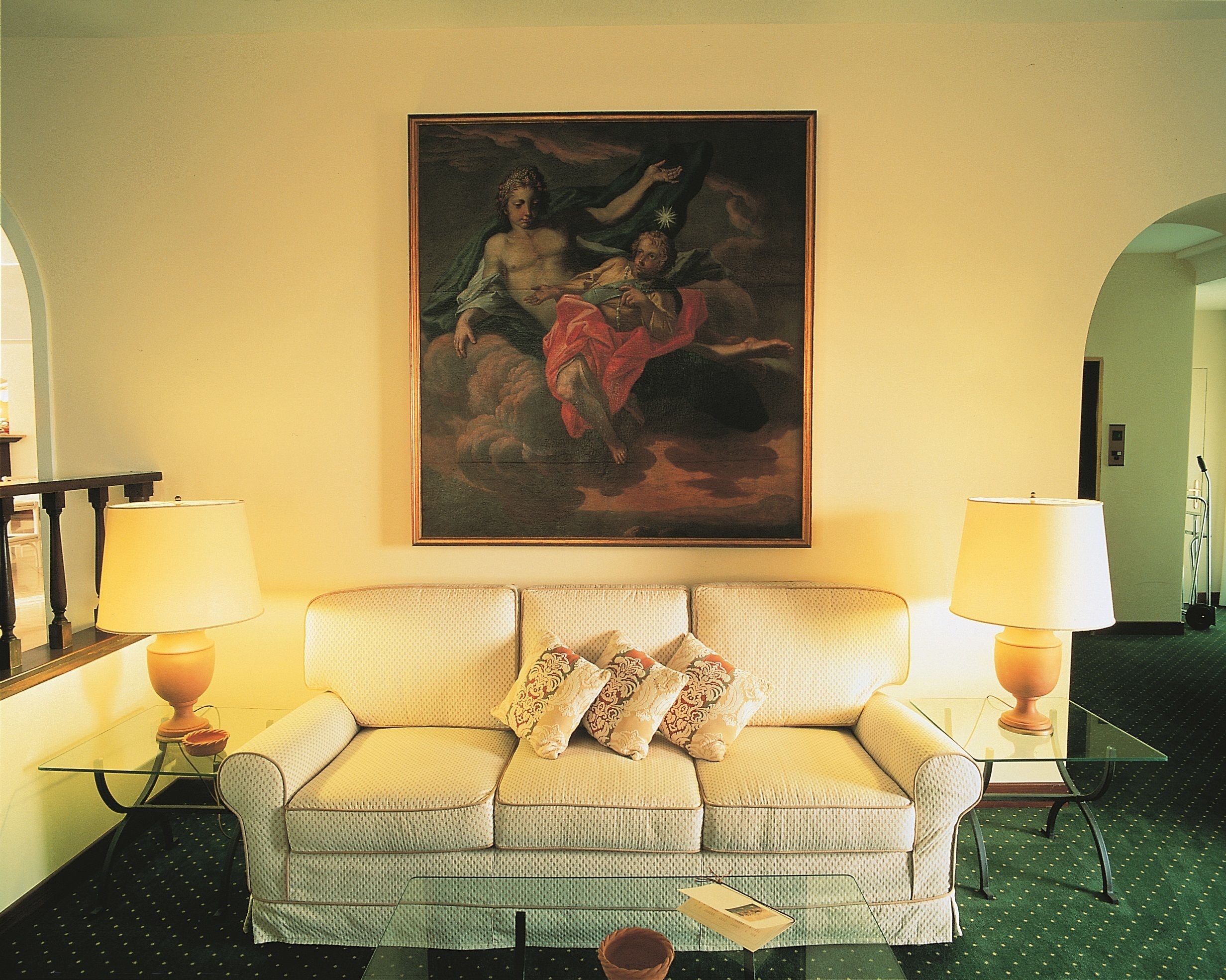 A hotel room with a beige sofa, two lamps, a framed painting, and a glass coffee table.