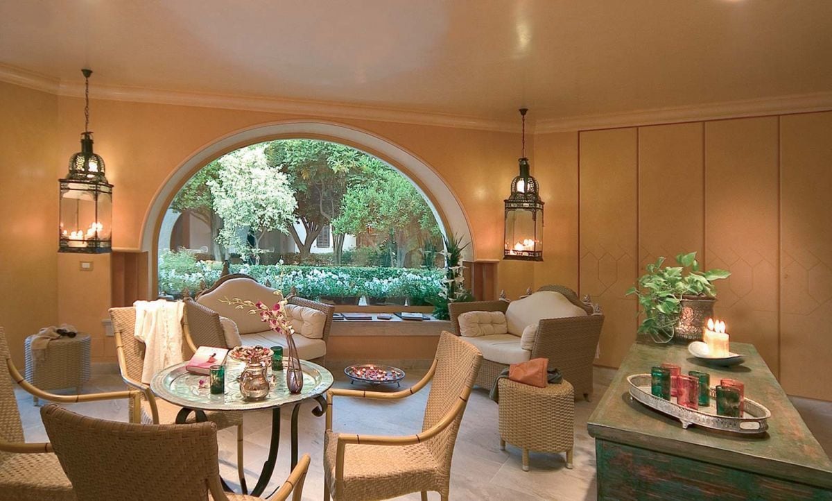 Un coin salon accueillant, avec des meubles en osier, une table ronde ornée d’un vase et une vue sur un jardin, visible à travers une fenêtre en arc de cercle.