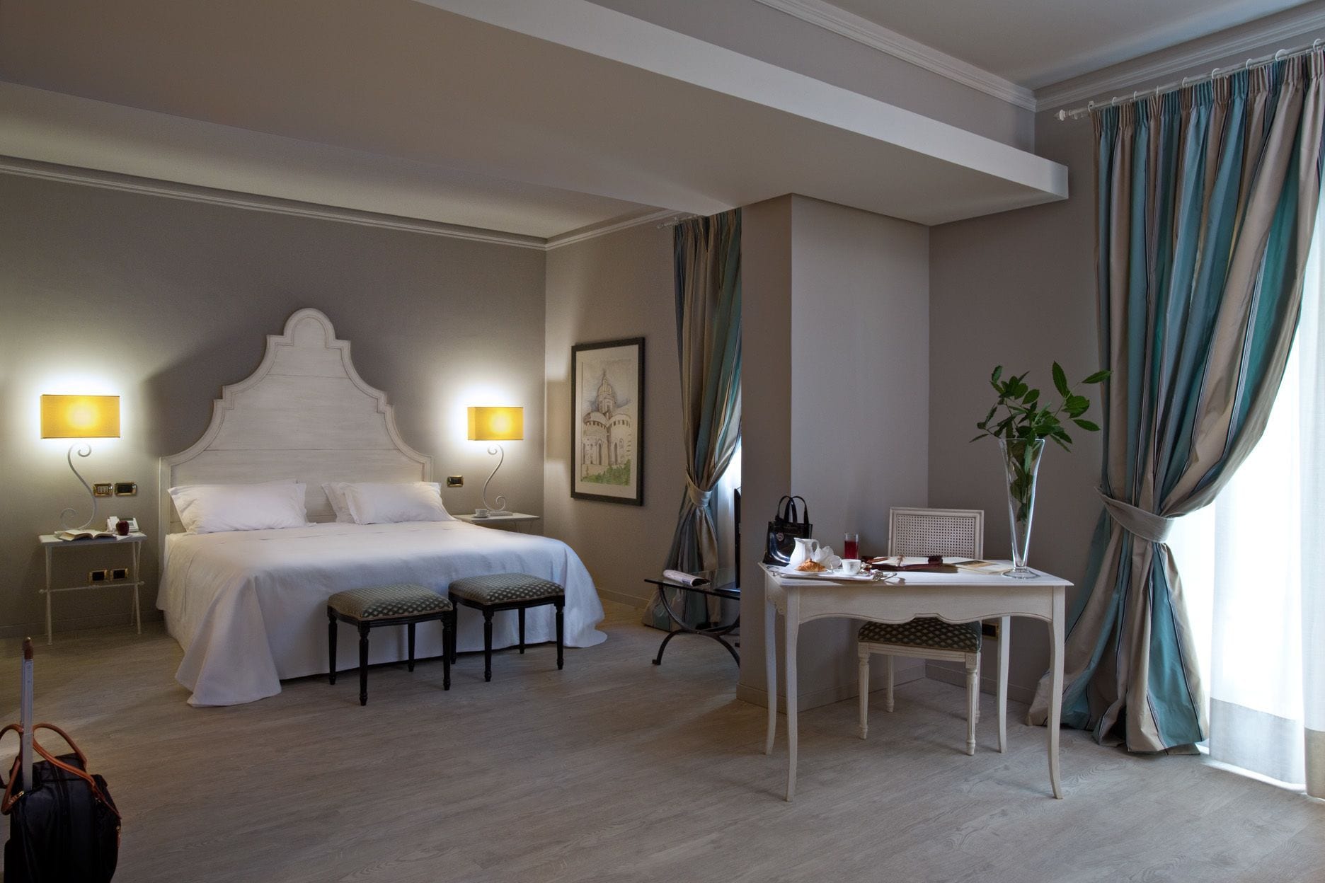Een hotelkamer met een groot bed, twee bedlampen, een klein tafeltje met een vaas, en een stoel.