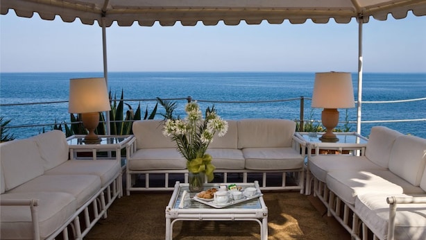 Un espacio de estar al aire libre cubierto con mobiliario blanco y lámparas, con vistas al mar.