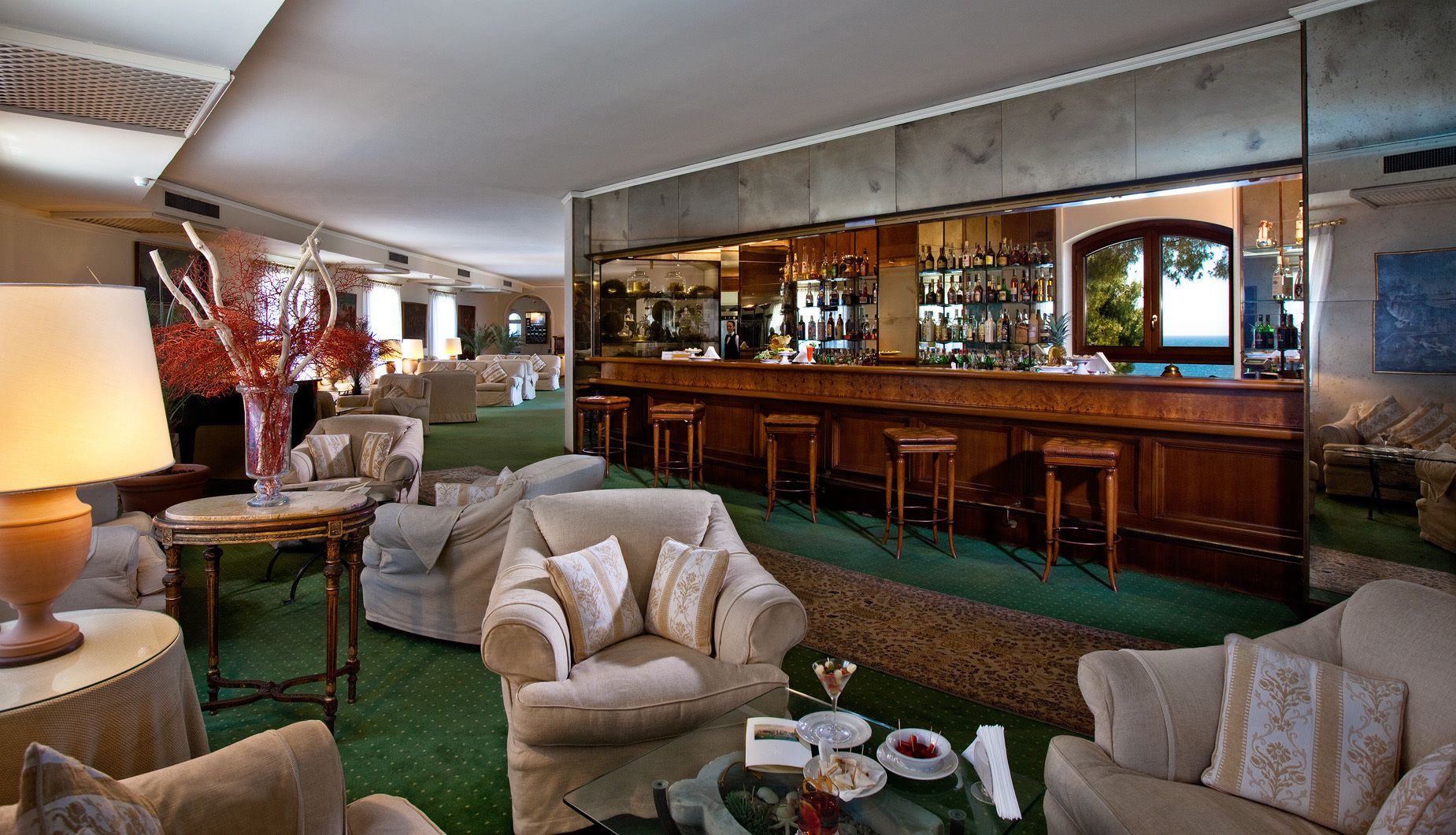 Un bar con mostrador de madera y taburetes, una zona de estar con sofás y sillones, y vista al mar a través de una ventana.