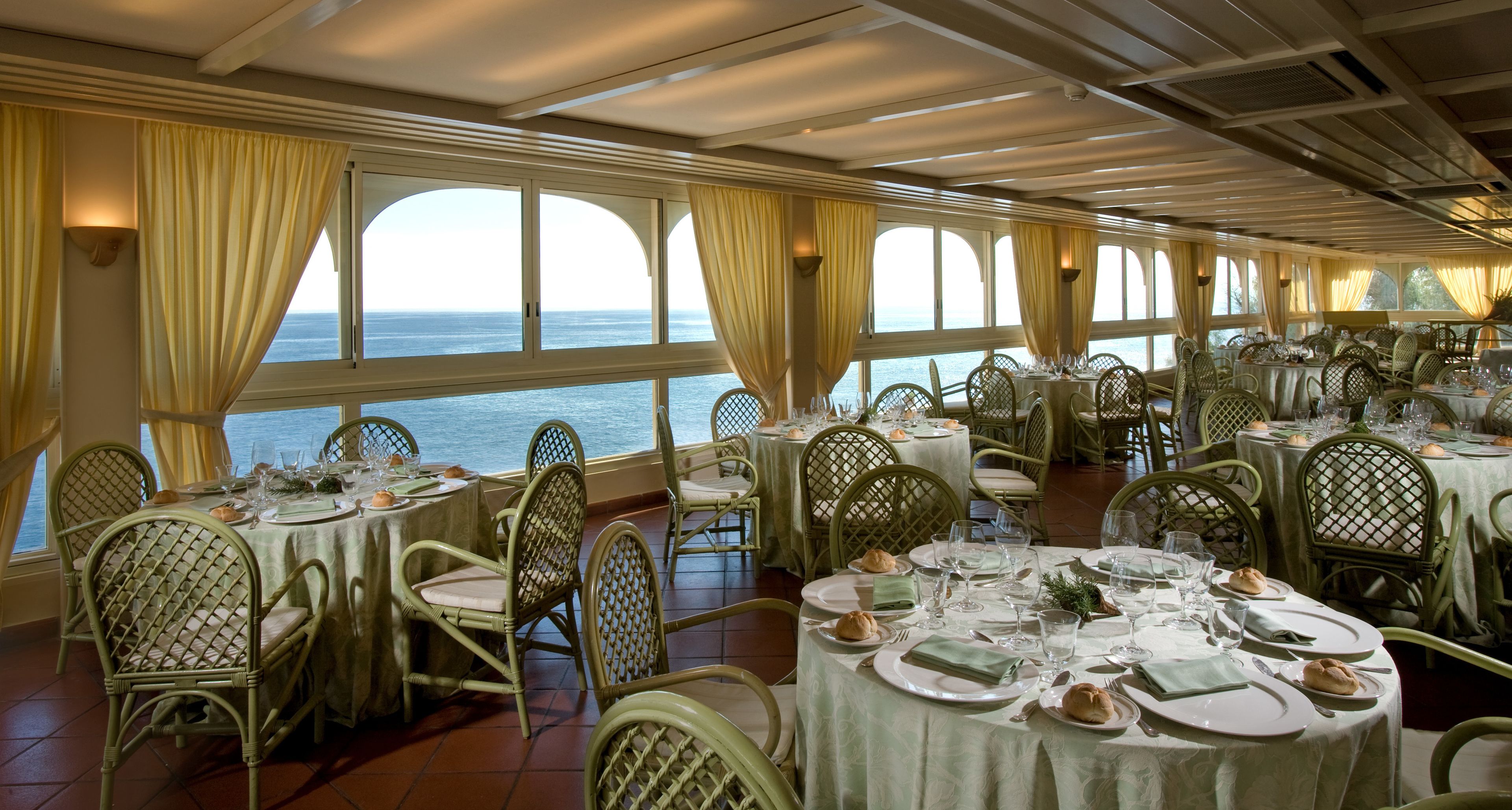 Un espace repas avec des tables dressées pour accueillir plusieurs convives, de grandes fenêtres avec des rideaux et une vue sur la mer.