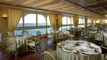 Un comedor con mesas dispuestas para varios comensales, ventanas amplias con cortinas y vistas al mar.