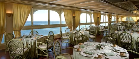 A dining area with tables set for multiple guests, large windows with curtains, and a view of the sea.