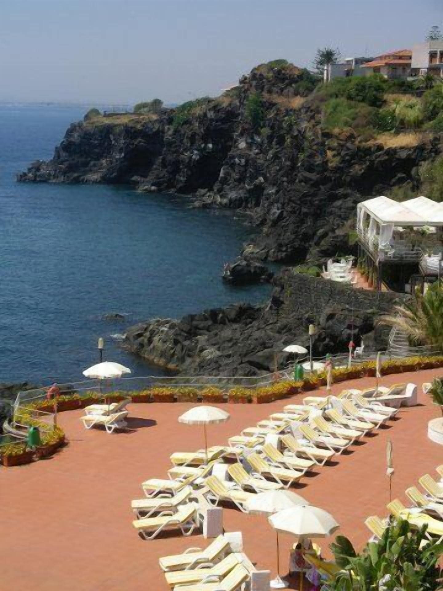 A coastal hotel with a terrace of sun loungers and umbrellas, overlooking the sea and rocky cliffs.