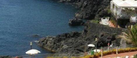 A coastal hotel with a terrace of sun loungers and umbrellas, overlooking the sea and rocky cliffs.