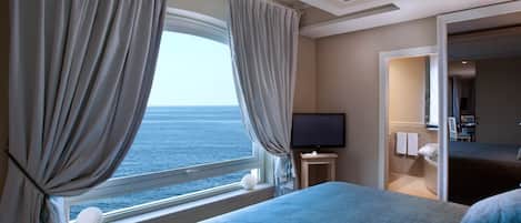 Habitación de hotel con una cama grande, un televisor sobre una mesita y vista al mar por la ventana.