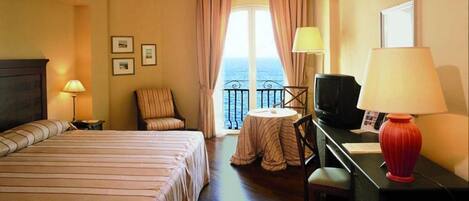 Een hotelkamer met een bed, een bureau met een televisie, een stoel, een kleine tafel en een balkon met gordijnen.