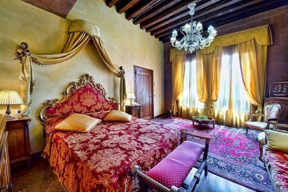 Ein Zimmer mit einem großen, aufwendig gestalteten Bett, einer Kristalllüster und einer klassischen Holzbalkendecke.