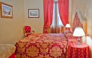 A hotel room with a large bed, a chair, a table with a lamp, and two framed pictures on the wall.