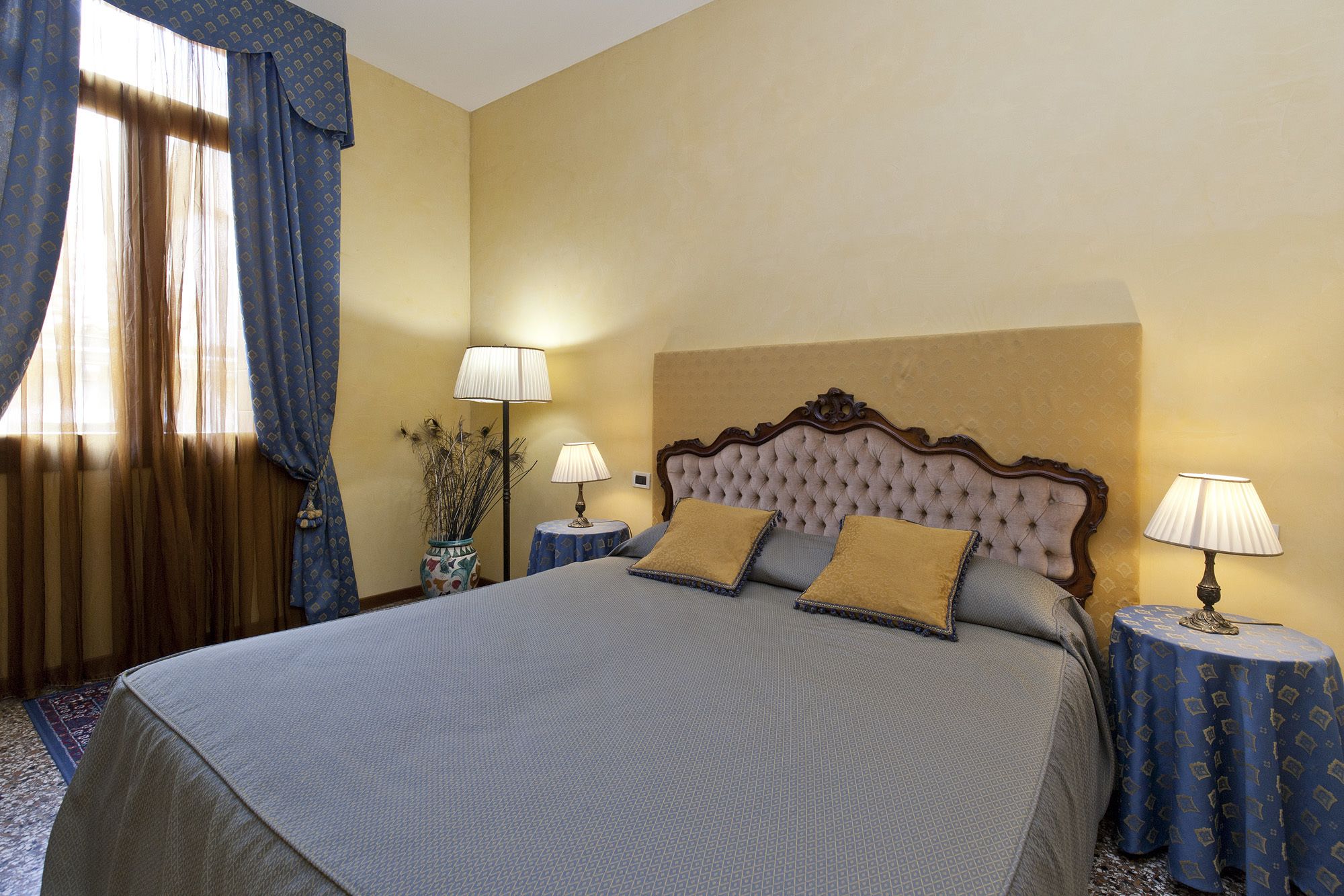 A neatly arranged hotel room with a large bed, two bedside tables with lamps, a vase with dried plants, and a window with blue curtains.