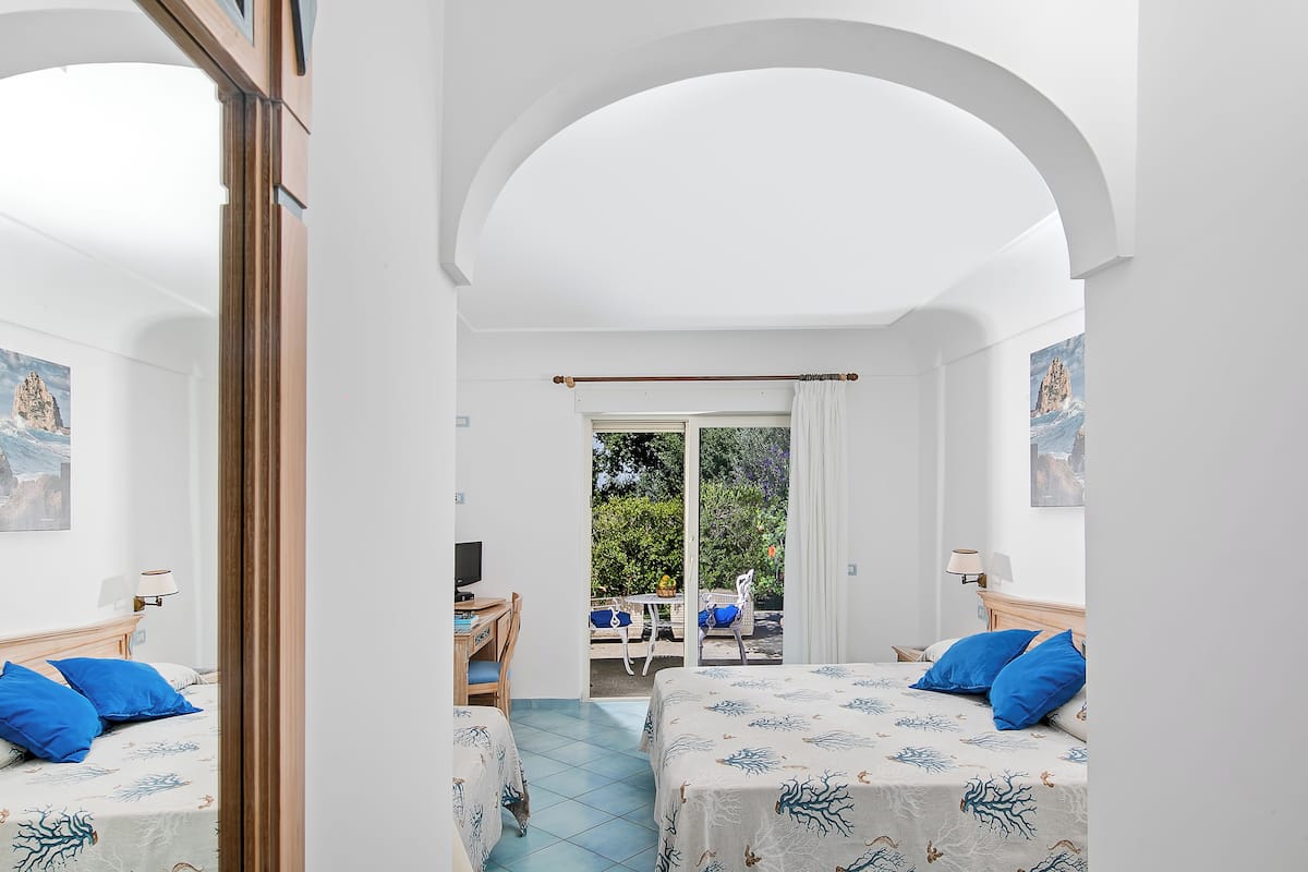 A room with two beds, a desk, and a balcony with outdoor seating.