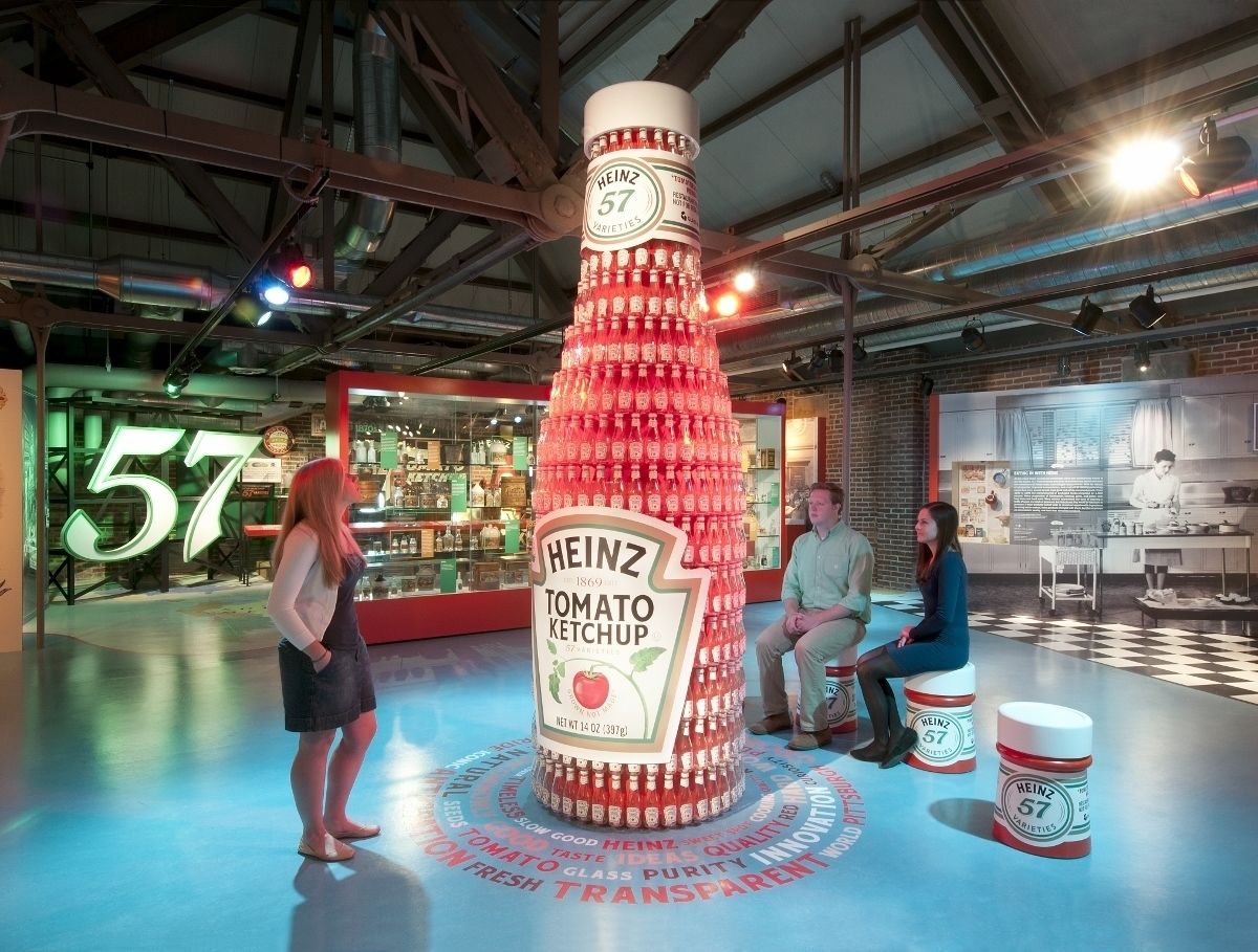 Uma grande escultura de garrafa de ketchup Heinz em um espaço interno moderno, com tetos aparentes e elementos industriais.