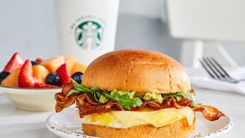Un sándwich de desayuno con tocino, lechuga y queso en un pan, un tazón de fruta y una taza de Starbucks.